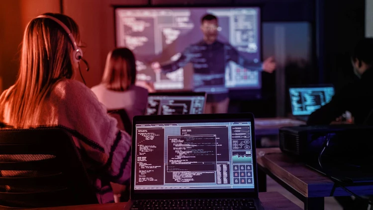 Group of software developers in training, with a female programmer in the foreground working on code on her laptop and a projected technical diagram in the background.